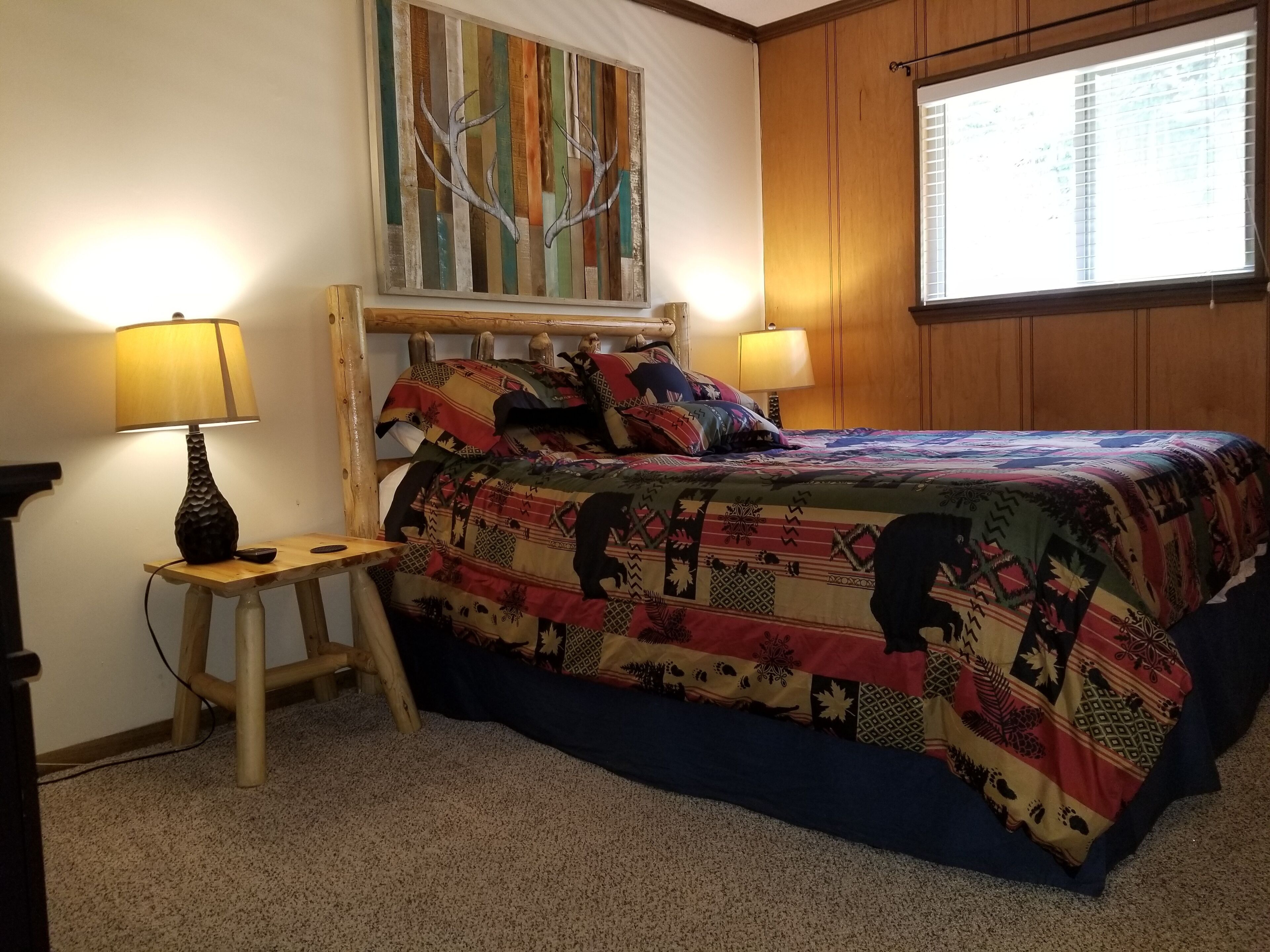 Une chambre à coucher avec une tête de lit en bois, une courtepointe aux couleurs vives, deux lampes de chevet, un petit tabouret et une fenêtre avec des stores.