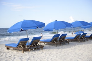 A row of wooden beach chairs with blue cushions and matching umbrellas on a sandy beach.