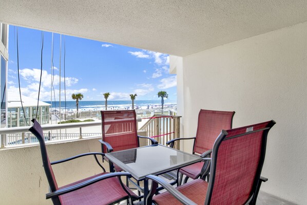 A balcony with a glass table and four chairs, overlooking a beach with palm trees and the ocean.