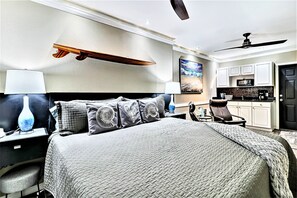 A hotel room with a large bed, a desk with a lamp, a chair, a ceiling fan, and a kitchenette.