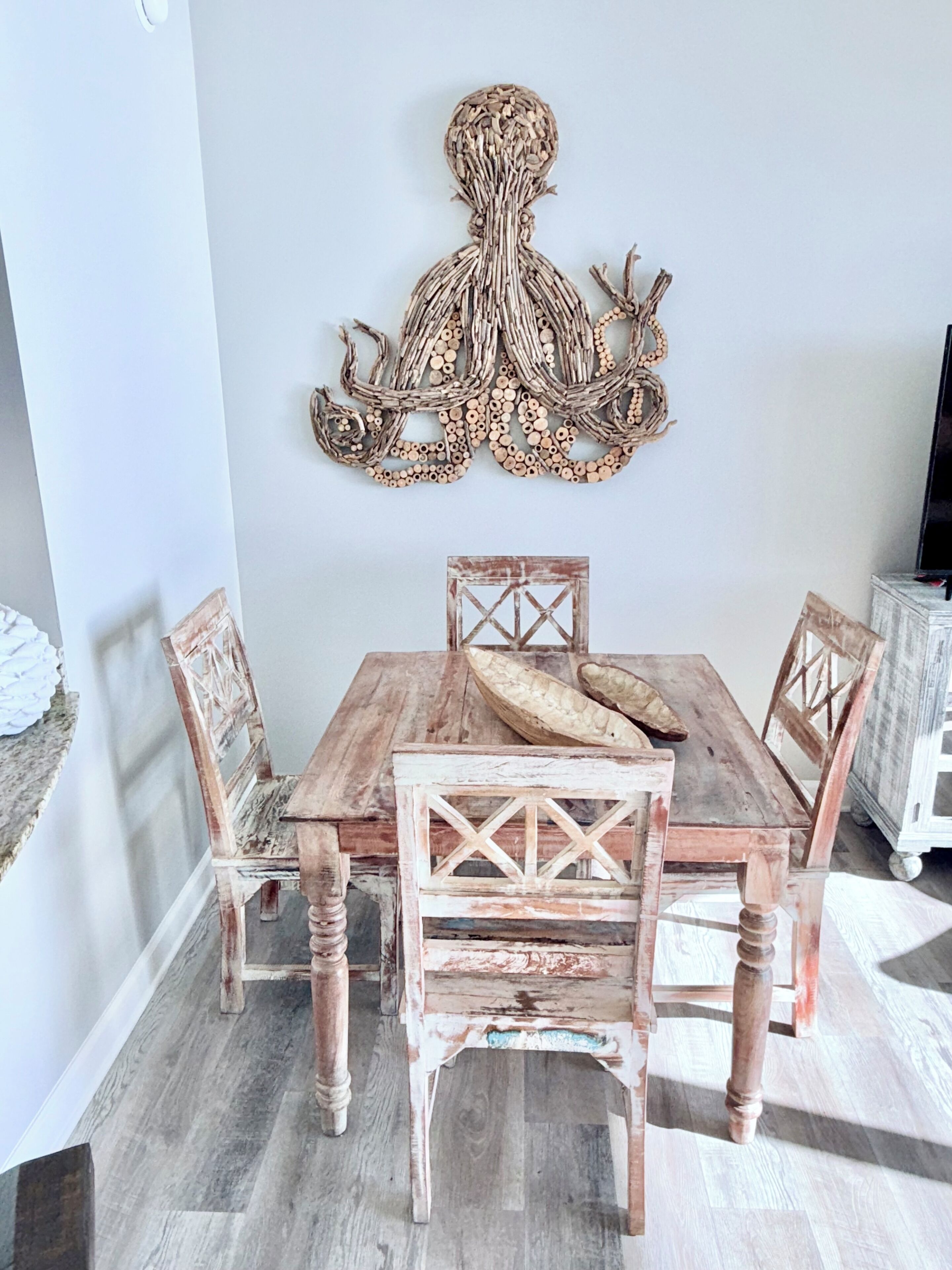 Uma mesa de jantar rústica de madeira com quatro cadeiras, um objeto decorativo na parede acima dela e uma televisão sobre um suporte à direita.