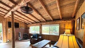 A rustic cabin interior with a wooden ceiling, a fireplace, a leather sofa, a table, and a lamp.