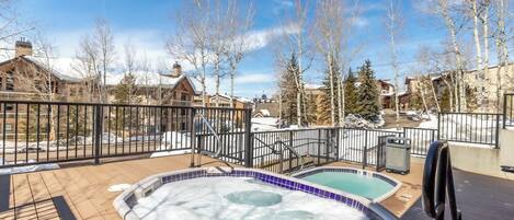 A hot tub area with a wooden deck and a pool, surrounded by a metal fence and leafless trees.