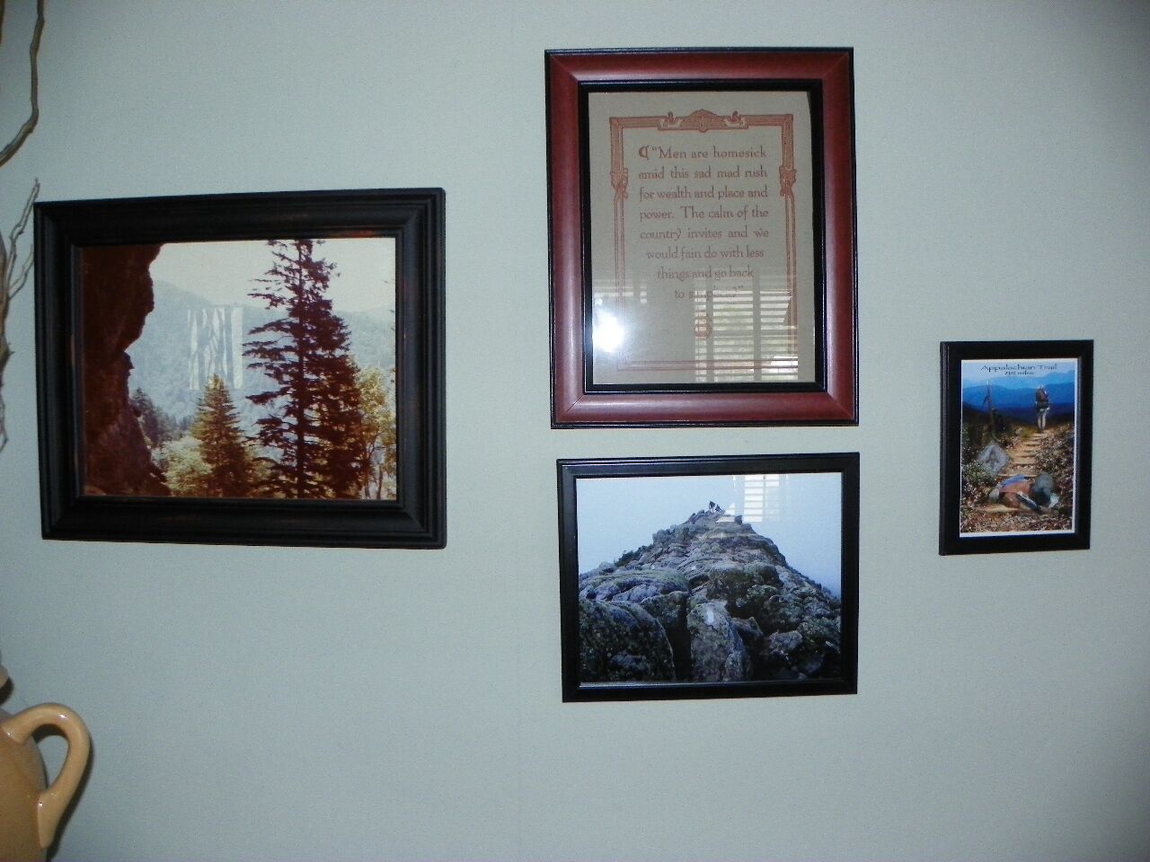 A wall with four framed pictures: a forest scene, a mountain peak, a framed quote, and a hiking trail.