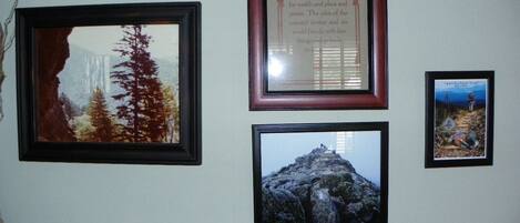A wall with four framed pictures: a forest scene, a mountain peak, a framed quote, and a hiking trail.