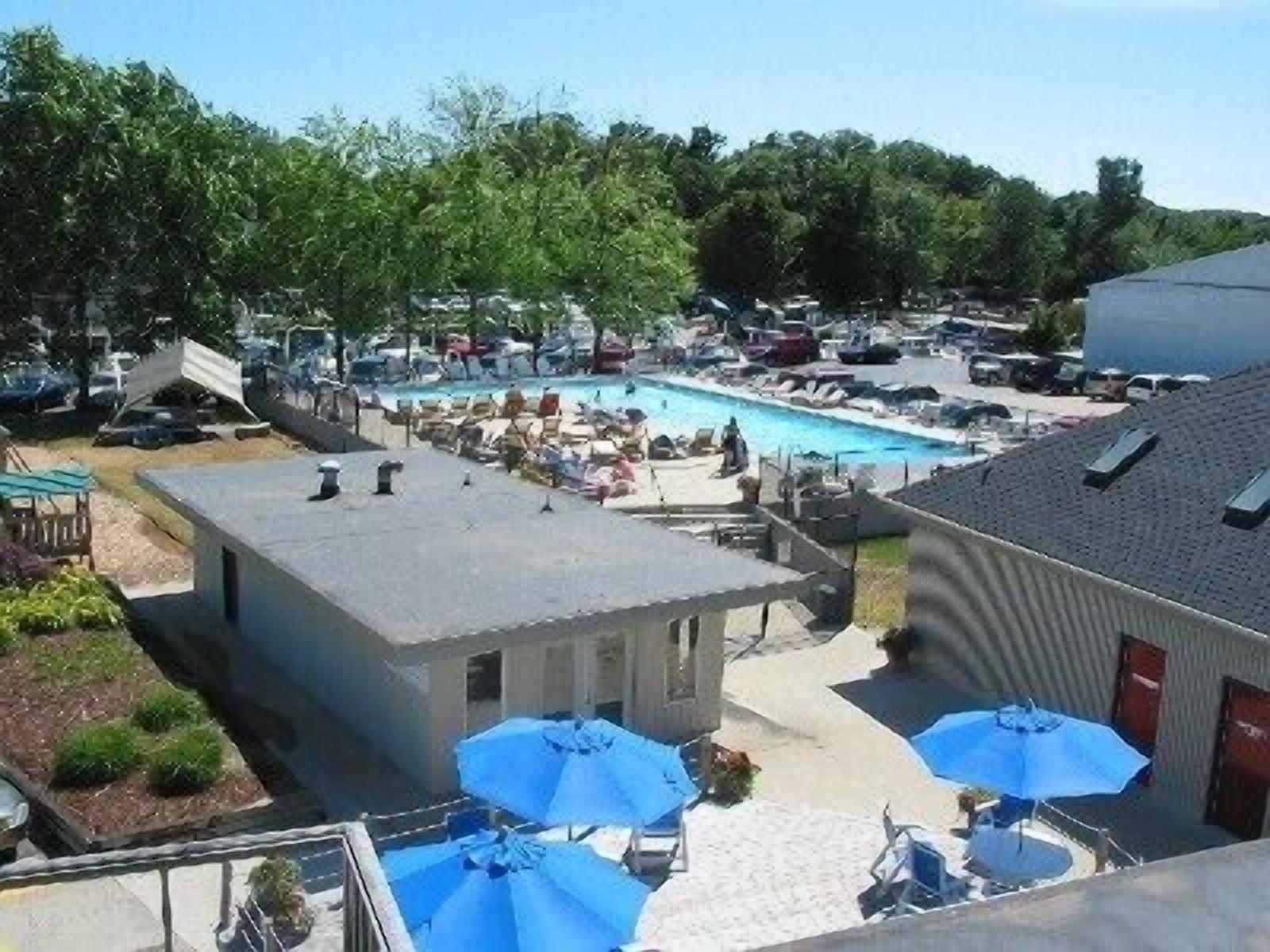 A pool area with lounge chairs and umbrellas, surrounded by parked cars and a building with a sloped roof.