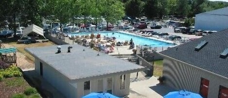 A pool area with lounge chairs and umbrellas, surrounded by parked cars and a building with a sloped roof.