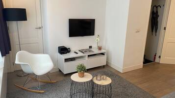 Een moderne woonkamer met een witte bank, een flatscreen televisie aan de muur, een kleine witte console tafel met een plant en een grijs, pluizig vloerkleed.