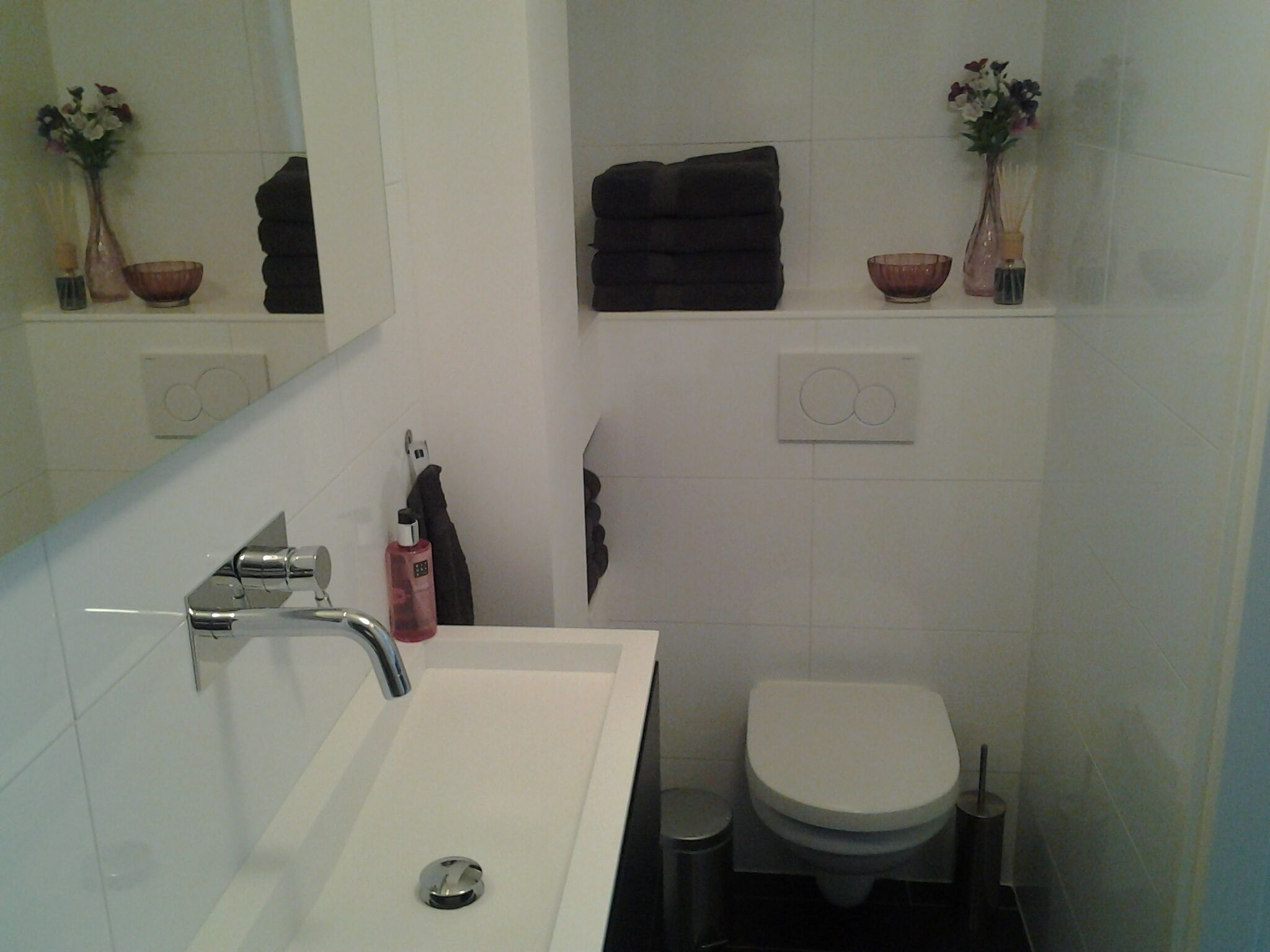 A bathroom with a white sink, a toilet, and a mirror. There are two flower vases on the wall, a towel hanging on a rack, and a soap dispenser on the sink.