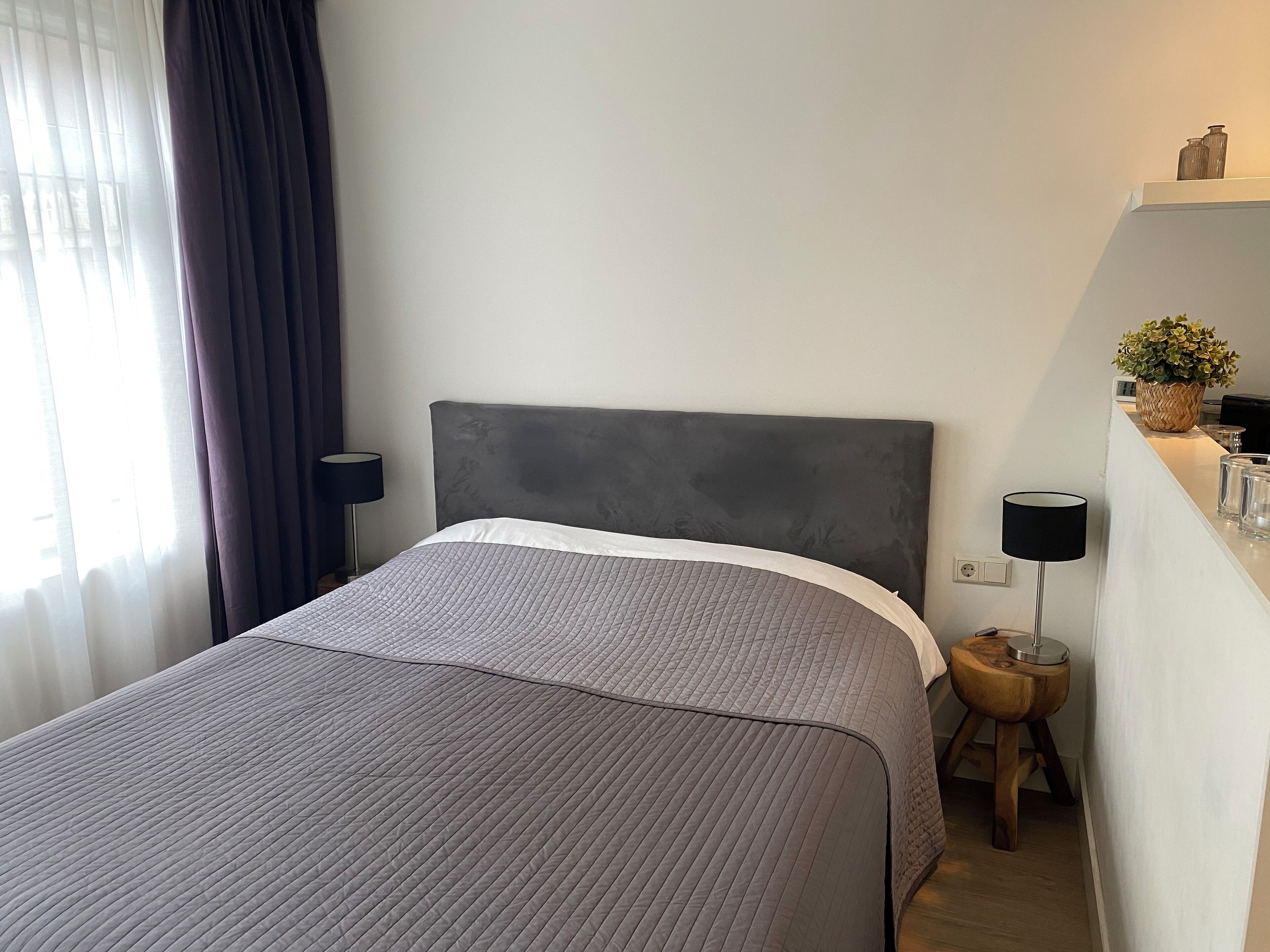Een netjes opgemaakt bed met een grijs hoofdbord en een grijze gewatteerde sprei. Er staat een klein houten krukje met een lamp naast het bed.