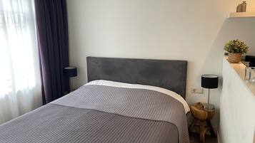 Een netjes opgemaakt bed met een grijs hoofdbord en een grijze gewatteerde sprei. Er staat een klein houten krukje met een lamp naast het bed.