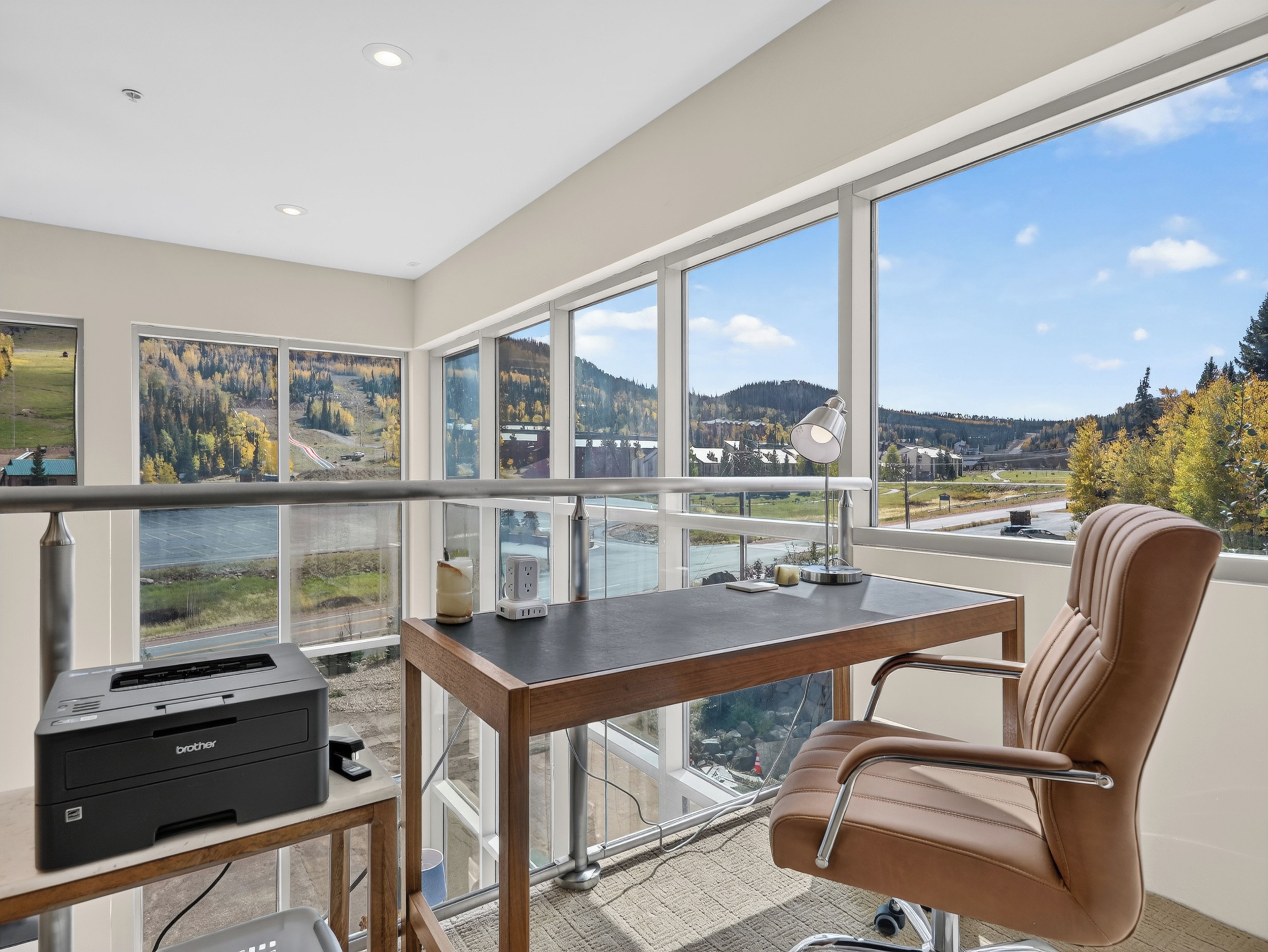 Un espace de bureau moderne comprenant un bureau, une chaise et une imprimante, avec vue sur un paysage pittoresque composé de montagnes et d’une rivière.