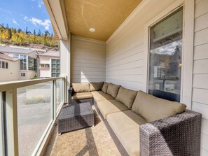 Un balcón con un sofá beige modular y sillas de mimbre, con vistas a una zona residencial con casas y árboles.