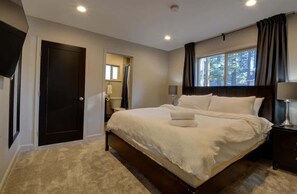 Quarto com uma cama grande, cabeceira escura, janela com cortinas, mesa de cabeceira e porta.