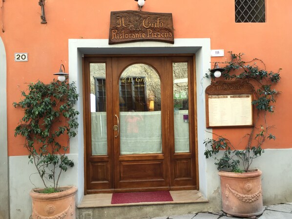 A wooden double door with a sign above it reading "Il Grifo Ristorante Pizzeria". There are two potted plants on either side of the door, and a red mat at the entrance. - Beauty And Serenity In A Premium Montalcino Location That Will Sleep 4 Adults. (Montalcino)