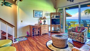A spacious living area with a wooden floor, a round wicker ottoman, a colorful sofa, and a television on a wooden stand. - Direct Oceanfront 2BD, Kaanapali/Kahana area, FANTASTIC! Home Away from Home! (Lahaina)