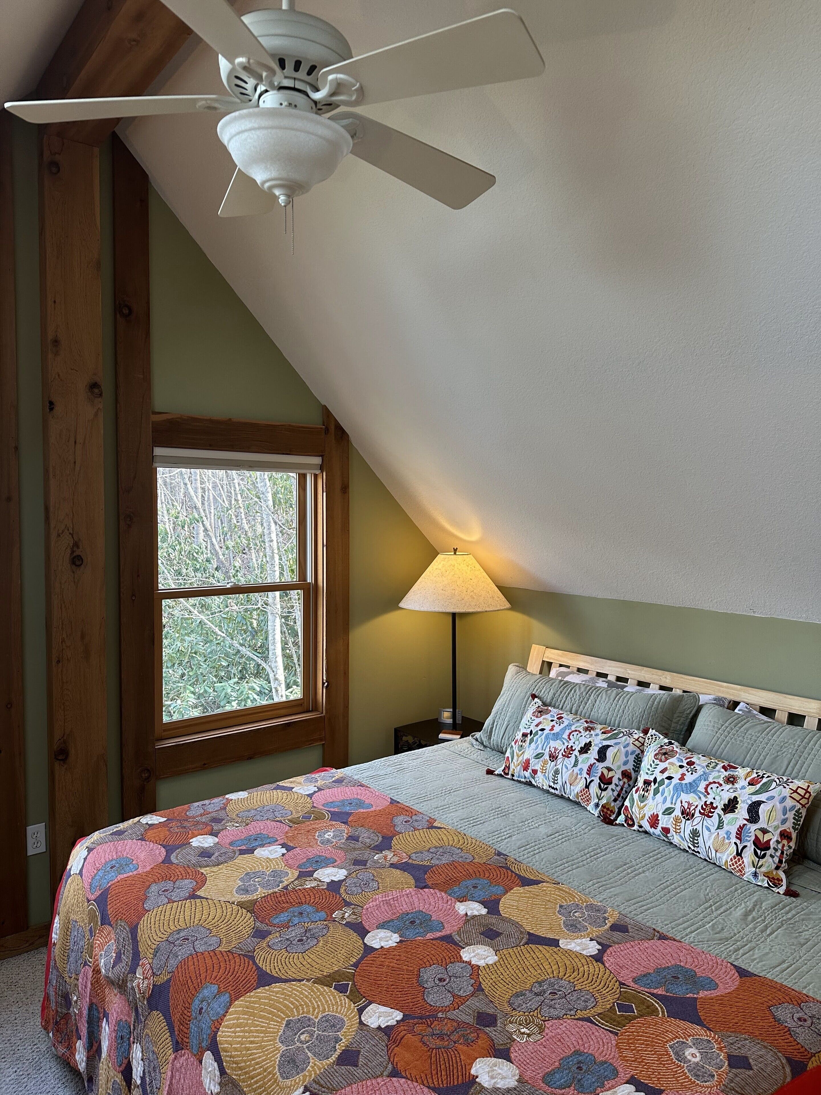 A bedroom with a sloped ceiling, a bed with a colorful quilt, a bedside lamp, and a window with a view of trees.