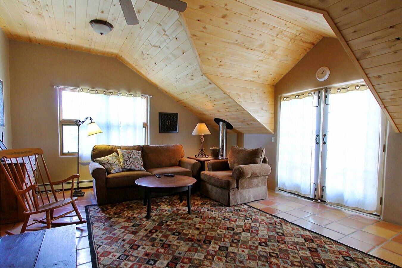 Un salon chaleureux avec un plafond en pente, une cheminée, un canapé, un fauteuil et un tapis à motifs.