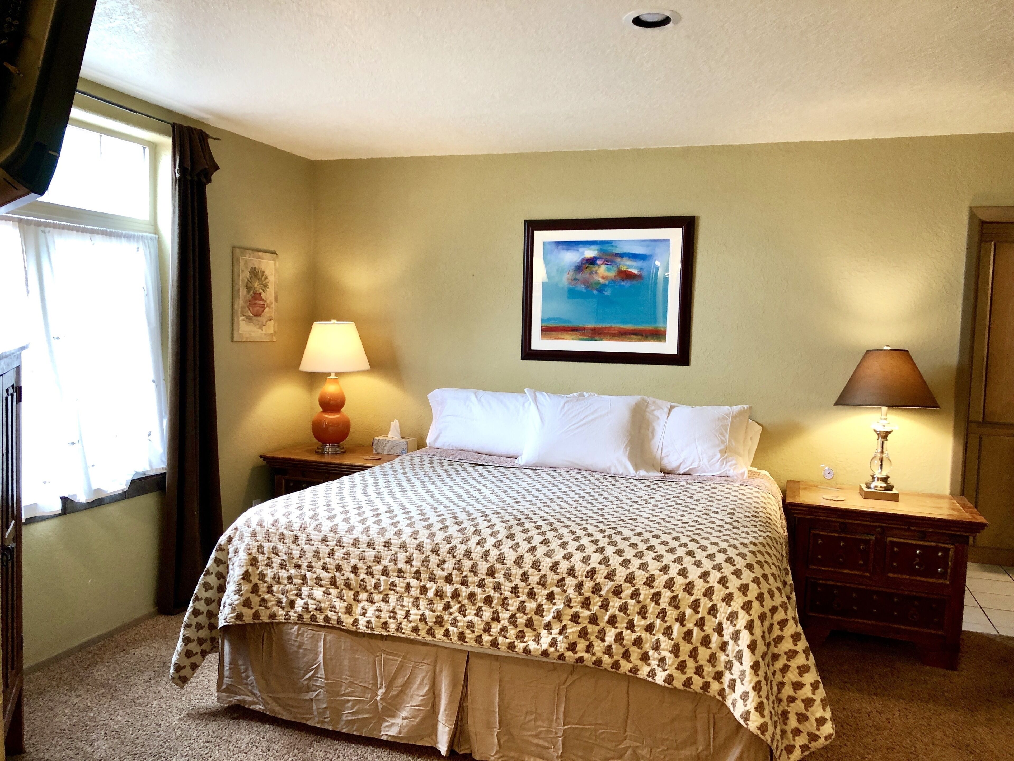 A hotel room with a bed, two bedside tables, a window with curtains, a painting on the wall, and a lamp on each table.