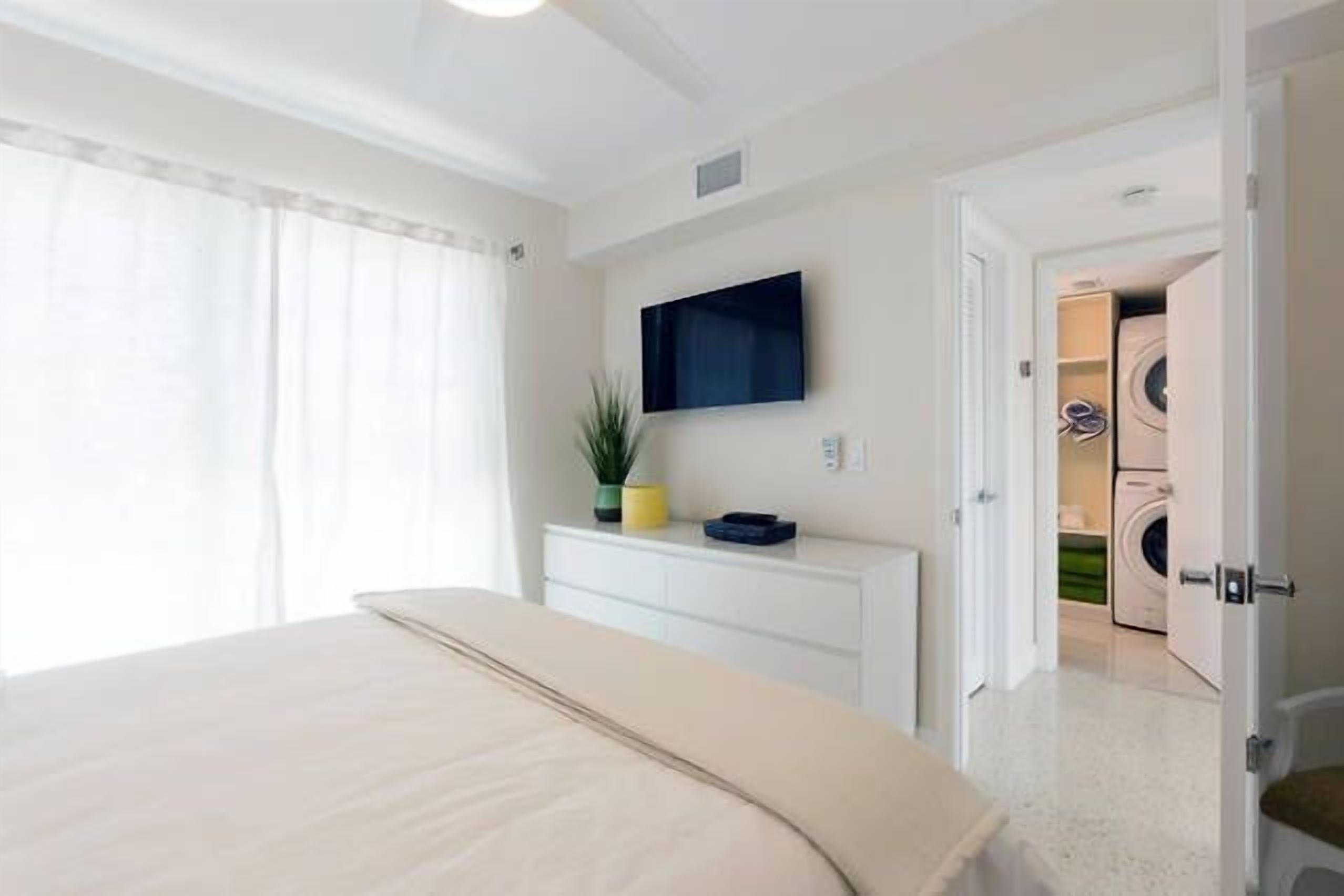 Une chambre à coucher avec un lit, un téléviseur fixé au mur et un coin buanderie visible à travers une porte ouverte.