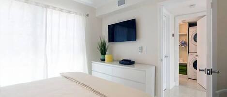 Une chambre à coucher avec un lit, un téléviseur fixé au mur et un coin buanderie visible à travers une porte ouverte.