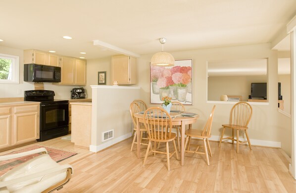 A modern kitchen with wooden cabinets, a black microwave, and a dining area with wooden chairs and a table.