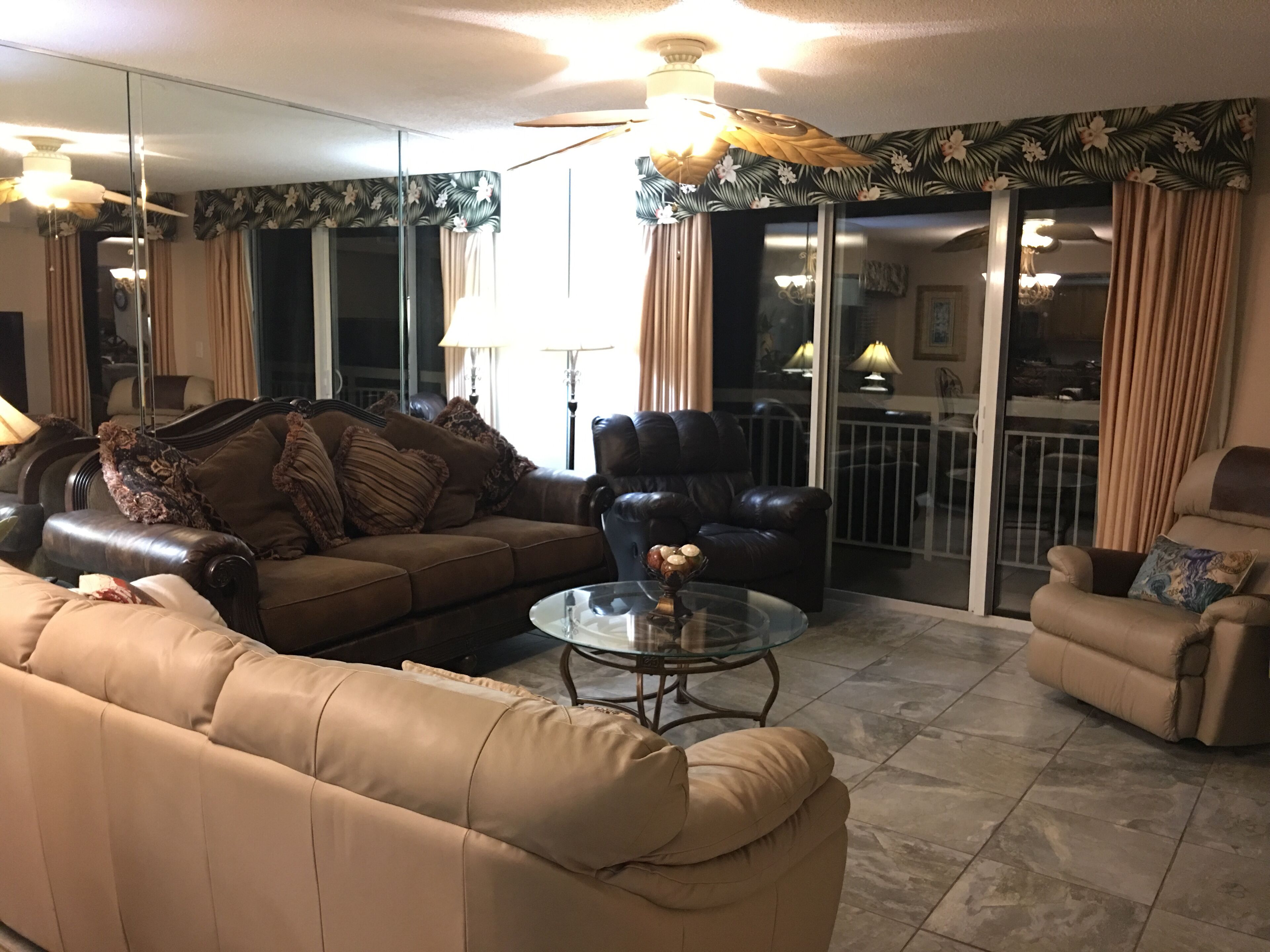 Uma sala de estar com um sofá de couro marrom, uma mesa de centro de vidro e uma poltrona de couro. Há uma porta de correr de vidro que dá para uma varanda.