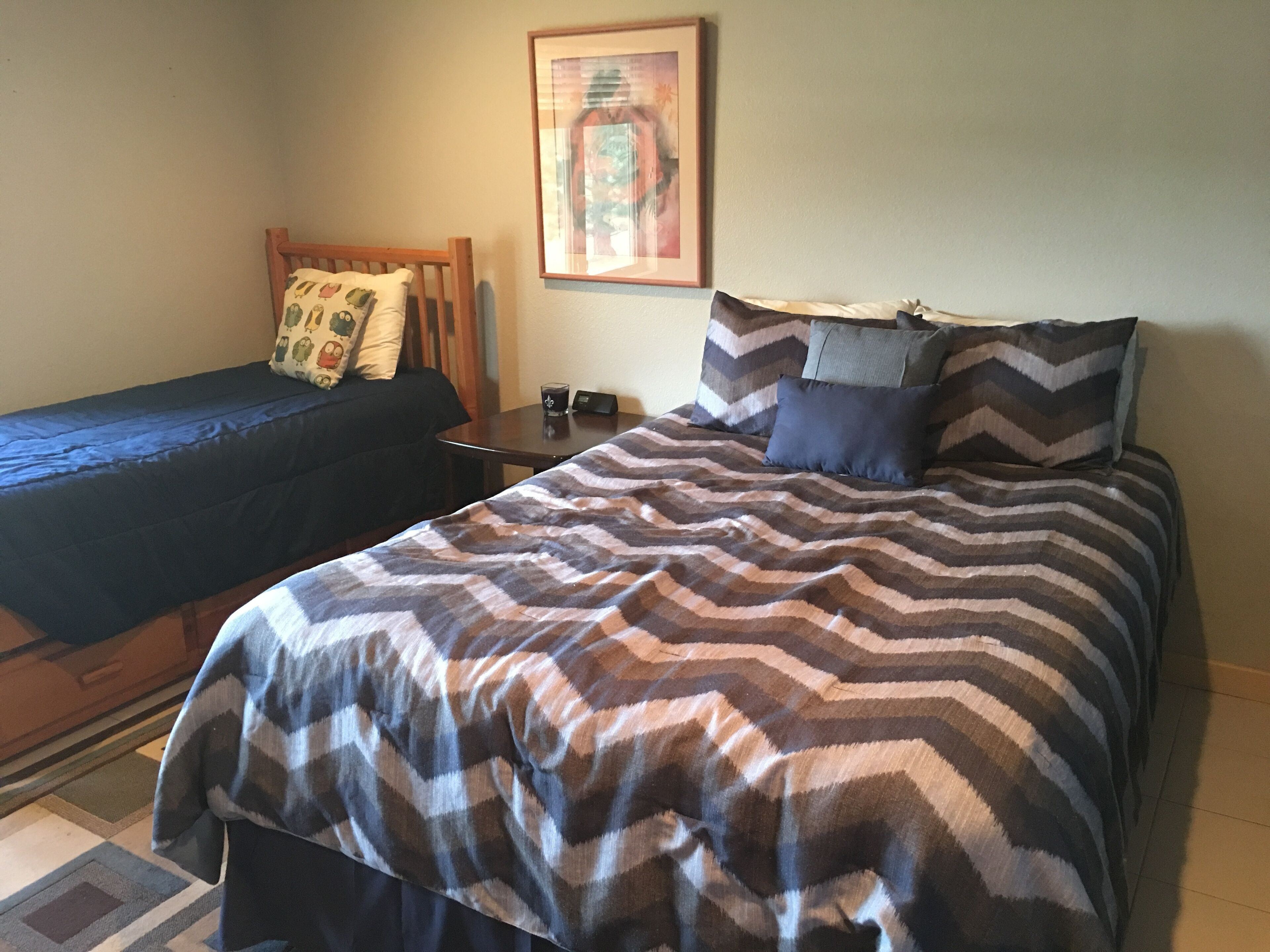 Een slaapkamer met een bed, een bijzetbed, een nachtkastje, een lijst met een foto aan de muur en een kamerkleed met dessin.