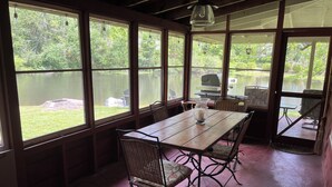 Un comedor con una mesa de madera y sillas de mimbre, ventanas amplias con vistas a un lago y follaje.