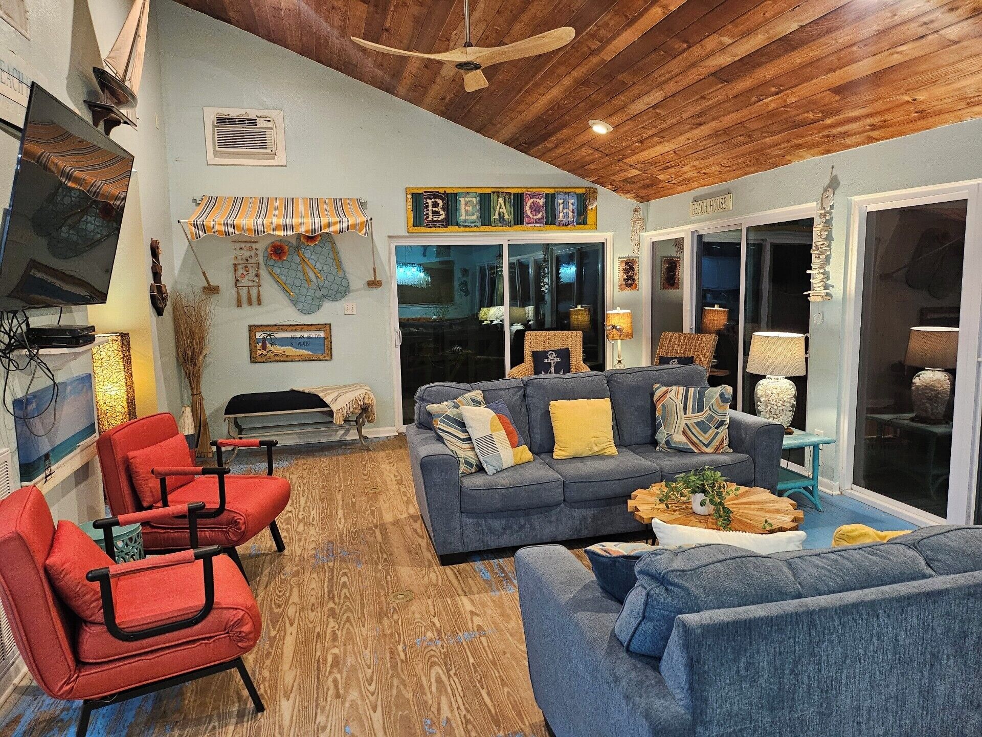 Un salon chaleureux avec un canapé bleu, des fauteuils rouges, un plafond en bois et une enseigne indiquant « Beach House ».