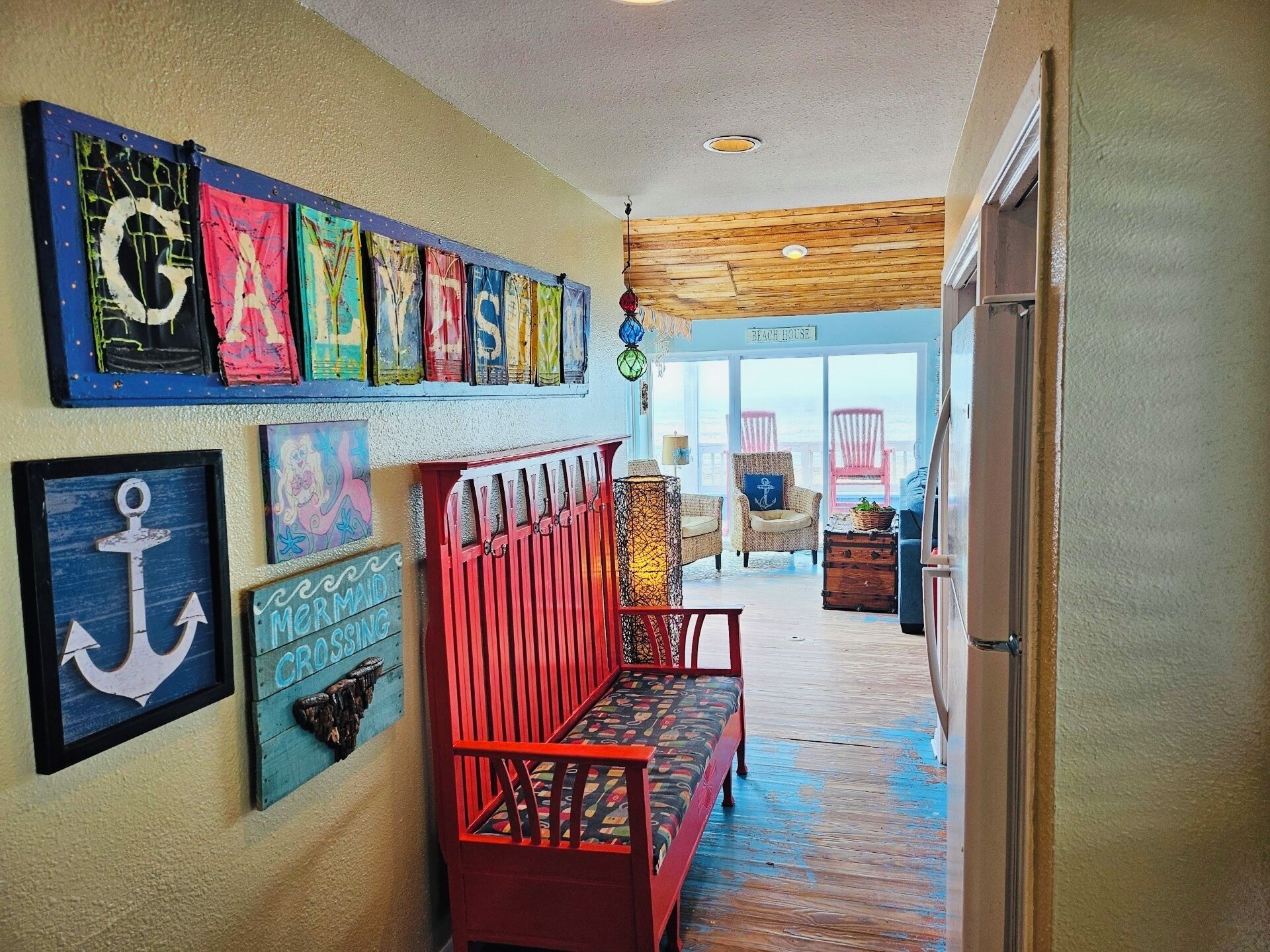 Un couloir avec un banc en bois, un mur orné d’œuvres d’art colorées et, à travers une porte vitrée, une vue sur une maison de plage.