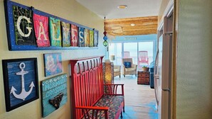 Un couloir avec un banc en bois, un mur orné d’œuvres d’art colorées et, à travers une porte vitrée, une vue sur une maison de plage.