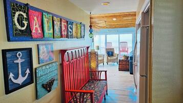 Un couloir avec un banc en bois, un mur orné d’œuvres d’art colorées et, à travers une porte vitrée, une vue sur une maison de plage.
