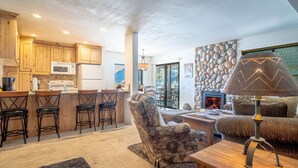 A spacious living area with a fireplace, a stone wall, and a kitchen with wooden cabinets and a bar area. - SV137 - Walk to Lifts & Town - Hot Tub & Pool (Ketchum)