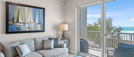 A living room with a sofa, a painting of sailboats, a lamp, and a view of the ocean through a balcony.
