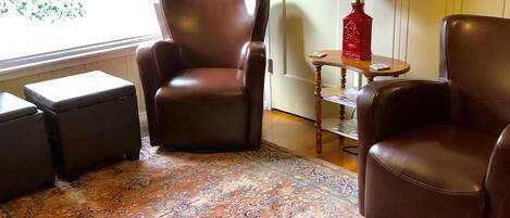 A living room with a patterned rug, two leather armchairs, a small table with a lamp, and a shelf with decorative items.