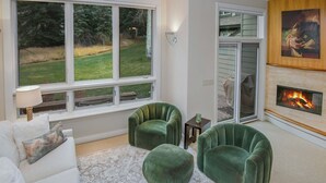 A living room with a white sofa, two green armchairs, a lace-patterned rug, a fireplace, and a large window with a view of a grassy yard. - Coldstream Townhome 38 | Stylish 3BR Cascade Village Retreat (Vail)
