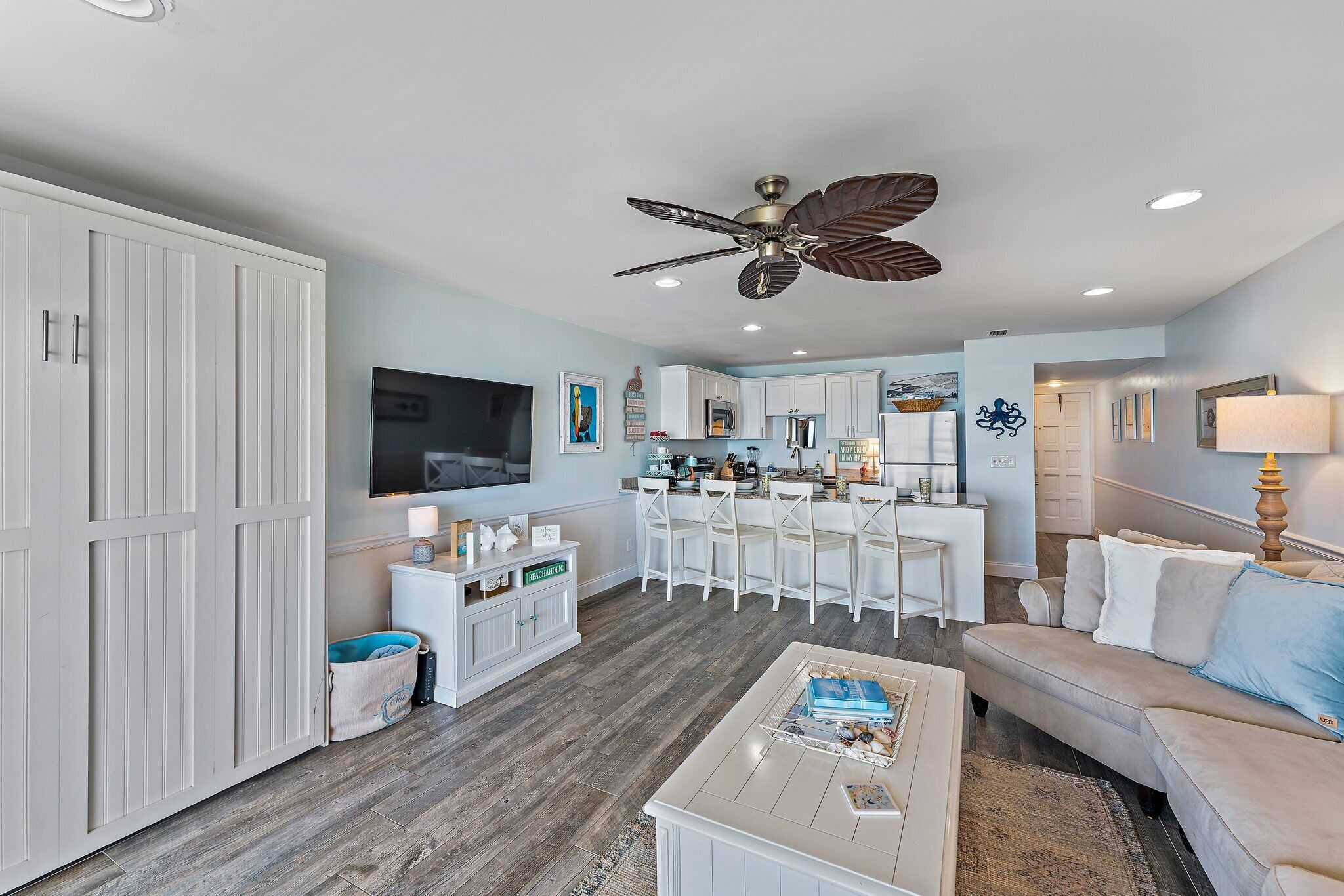 A modern living room with a large white cabinet, a ceiling fan, a flat-screen TV, and a kitchen area with white cabinets and a bar.