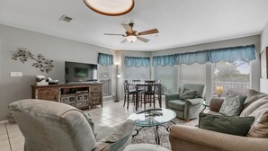 A living room with a TV, dining table, and chairs. There is a ceiling fan and a window with curtains. - Treetop Retreat on the Guadalupe Ultra Private 3rd Floor End Unit (New Braunfels)