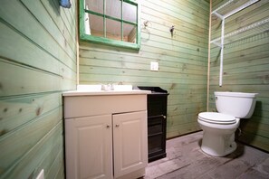 A small, basic bathroom with wooden walls, a white sink, a black trash bin, and a white toilet.