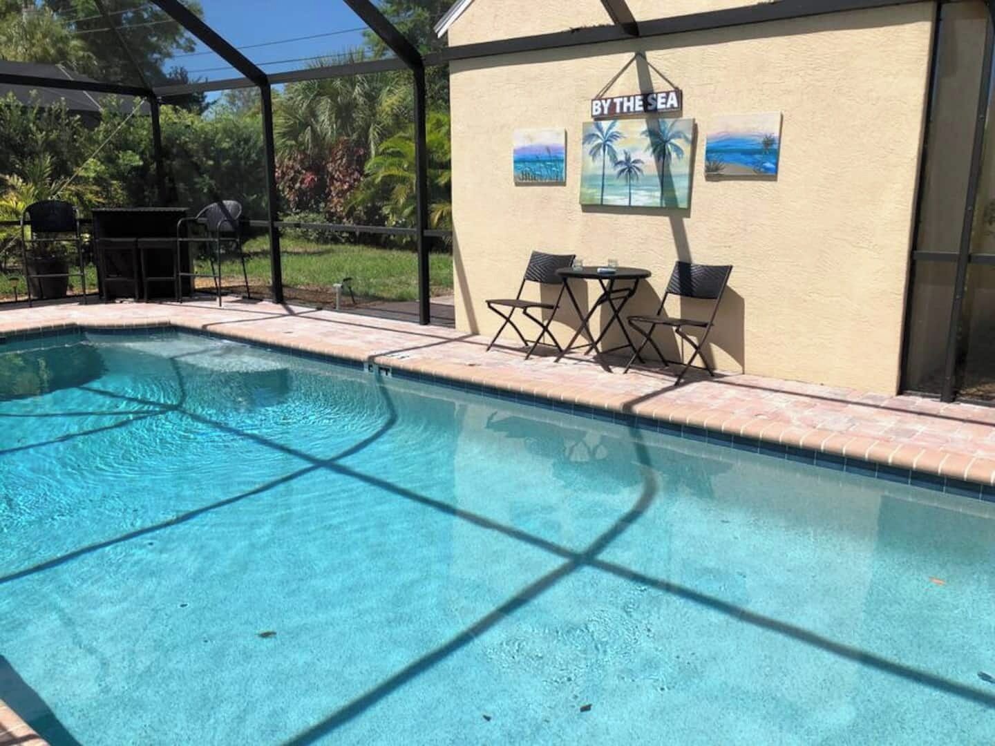 Une piscine dotée d’un espace couvert, une table avec des chaises et un mur orné d’une inscription « AU BORD DE LA MER » et de peintures représentant des palmiers.