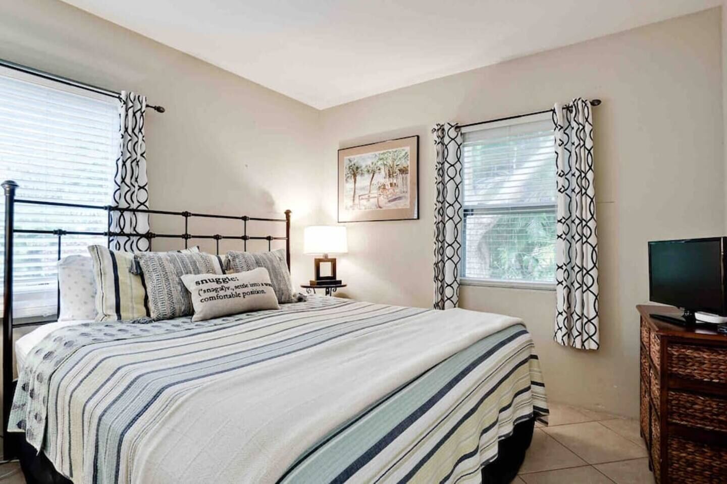 A bedroom with a bed, a bedside table with a lamp, a television on a stand, and patterned curtains.