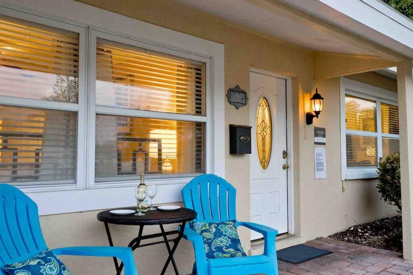 A building entrance with a door, two windows with blinds, a small table with a bottle and glasses, and two blue chairs.