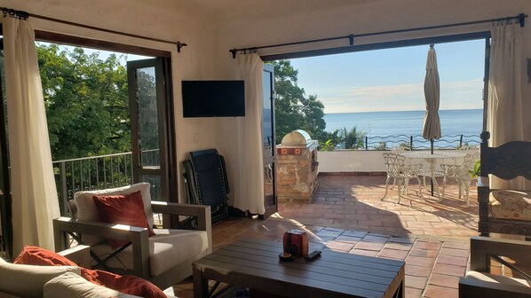 Een balkon met uitzicht op zee, een tafel, stoelen en een aan de muur bevestigde televisie.