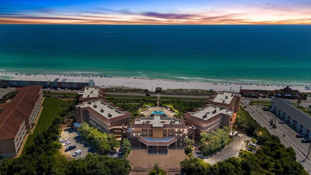 Luchtbeeld van een hotel met een centraal gelegen zwembad, omringd door weelderige begroeiing en een strand.