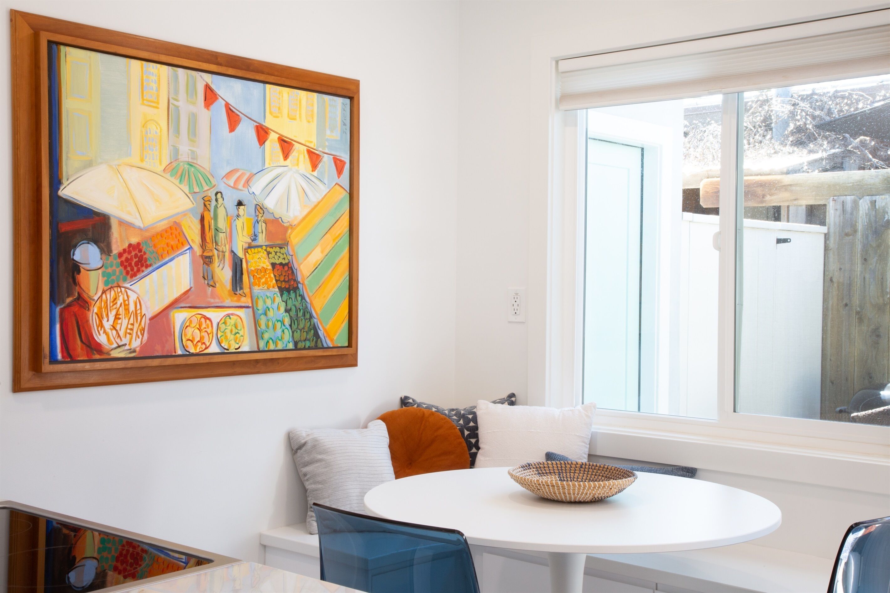 A dining area with a round table, a colorful abstract painting, and a view of the outdoors through a window.