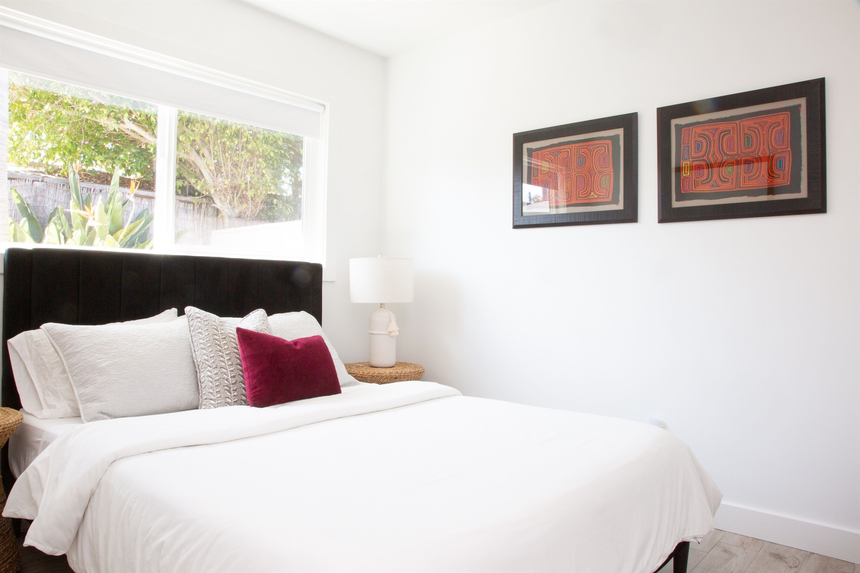 A bedroom with a large bed, white bedding, a black headboard, two framed artworks on the wall, a bedside lamp, and a view of the outdoors through a window.