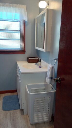Un baño con un lavabo blanco, un espejo encima, una ventana con persianas, un caja de madera sobre la encimera y una cesta de ropa.