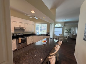 A modern kitchen with white cabinets, a black granite countertop, and a built-in oven. There is a dining area with wicker chairs and a view of the outdoors.