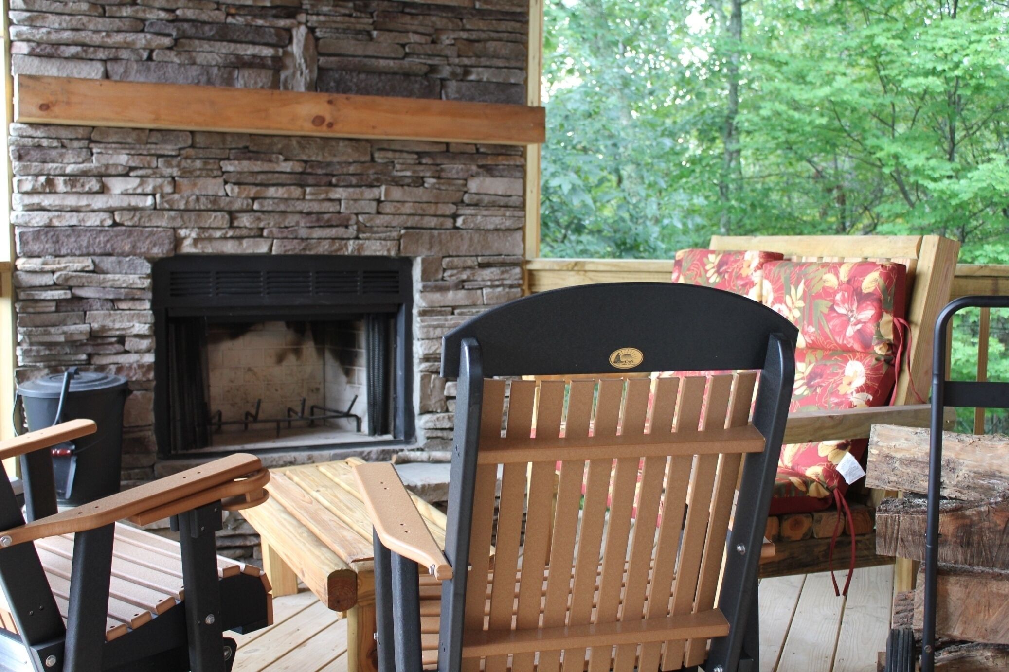 A stone fireplace with a log fire, wooden chairs, and a table in a covered outdoor area with a view of trees.